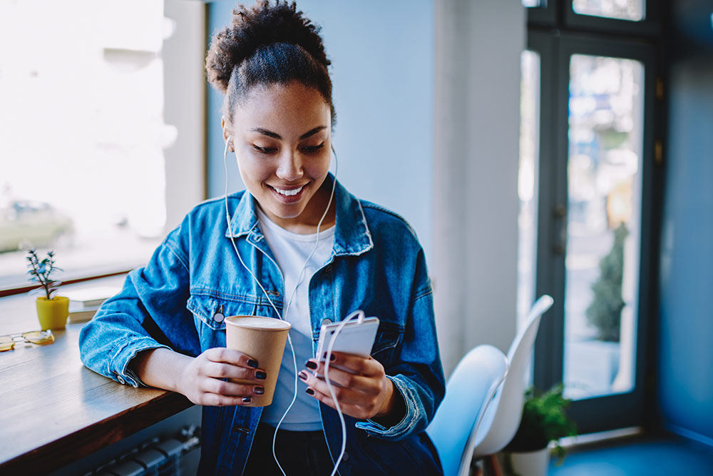 Woman sitting in coffee shop holding coffee while holding smartphone with headphones in, smiling at phone